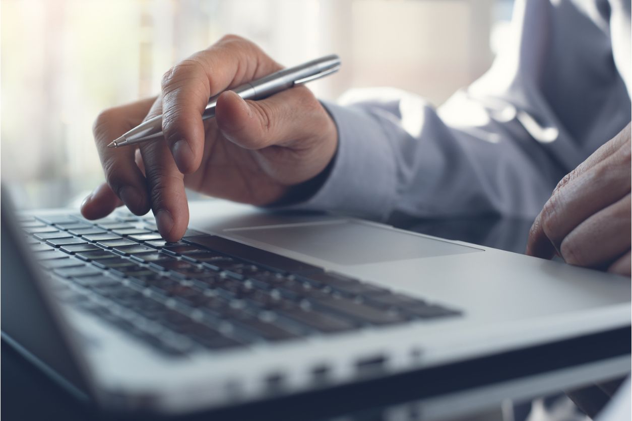 Meeting report in progress. Business man with pen in hand typing on laptop computer keyboard on desk, working in modern office with blurry background, close up.